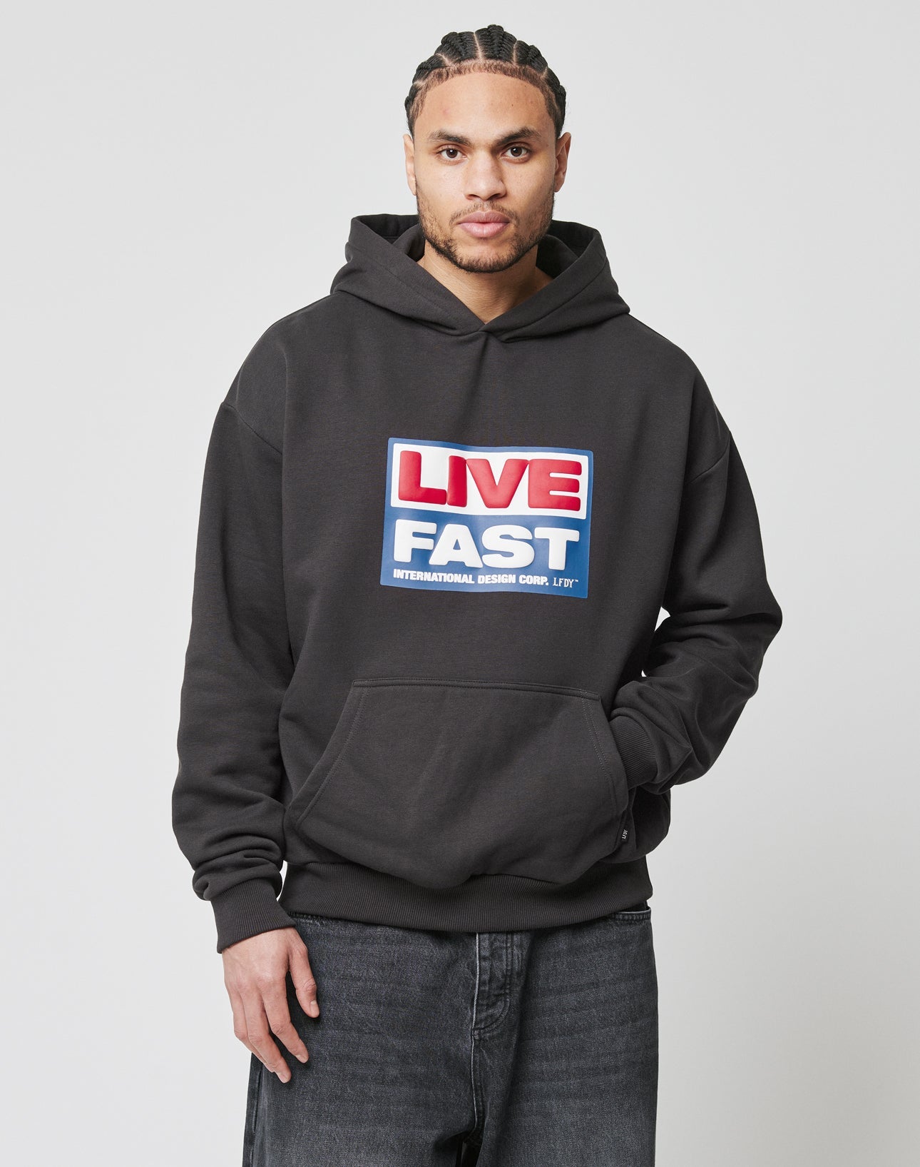 A man with braided hair wears the LIVE FAST DIE YOUNG Live Fast Box Hooded, an oversized black hoodie featuring a large front pocket and bold red, white, and blue puffer print, standing against a plain white background.