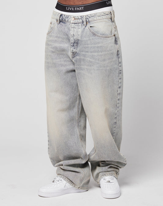 Wearing LFDY Baggy Denim by LIVE FAST DIE YOUNG with a wide straight leg, white sneakers, and a black LIVE FAST waistband, the cropped photo captures the lower body against a plain background.