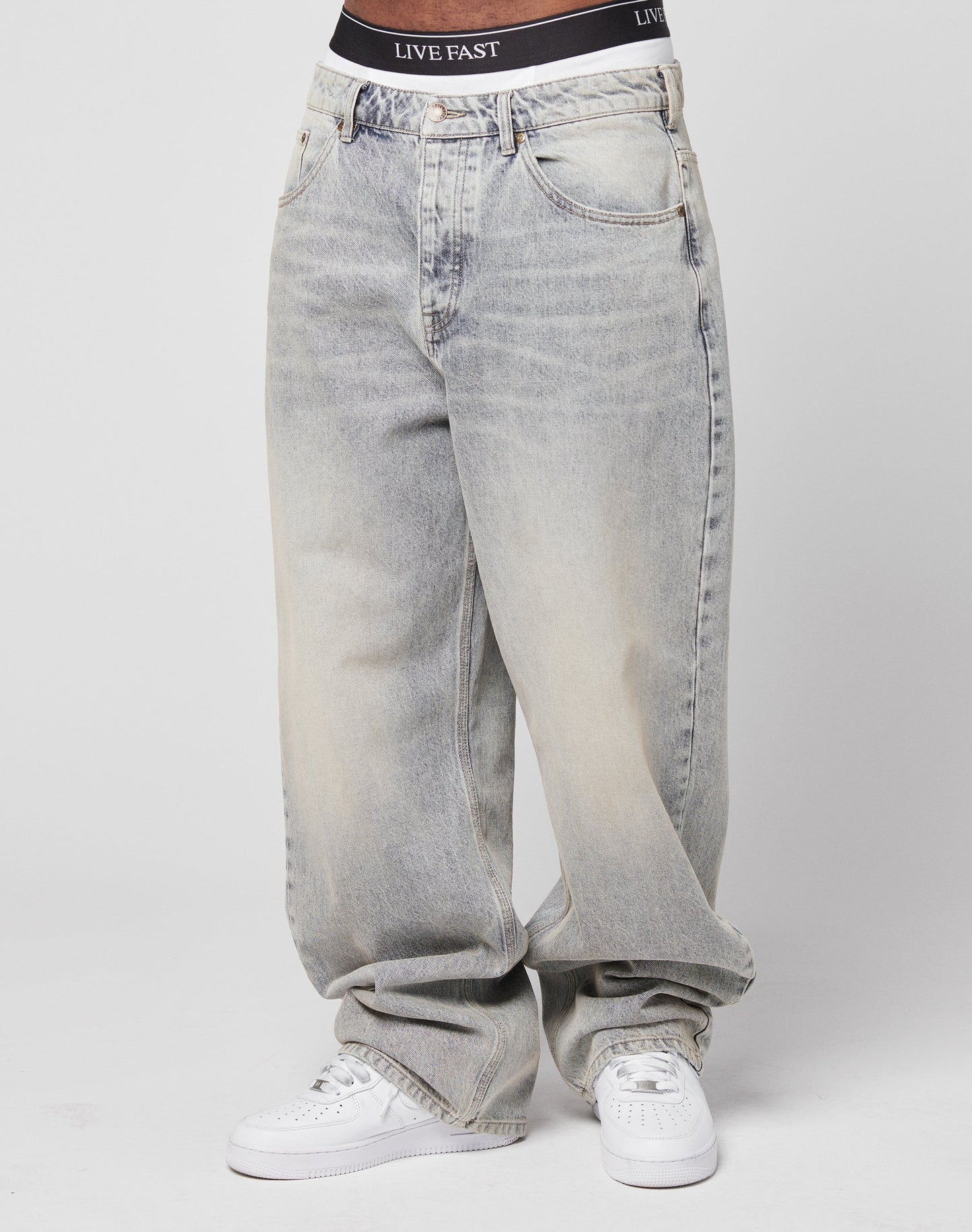 A person wearing LFDY Baggy Denim by LIVE FAST DIE YOUNG with a flag label, white sneakers, and black LIVE FAST waistband underwear stands against a plain white background.
