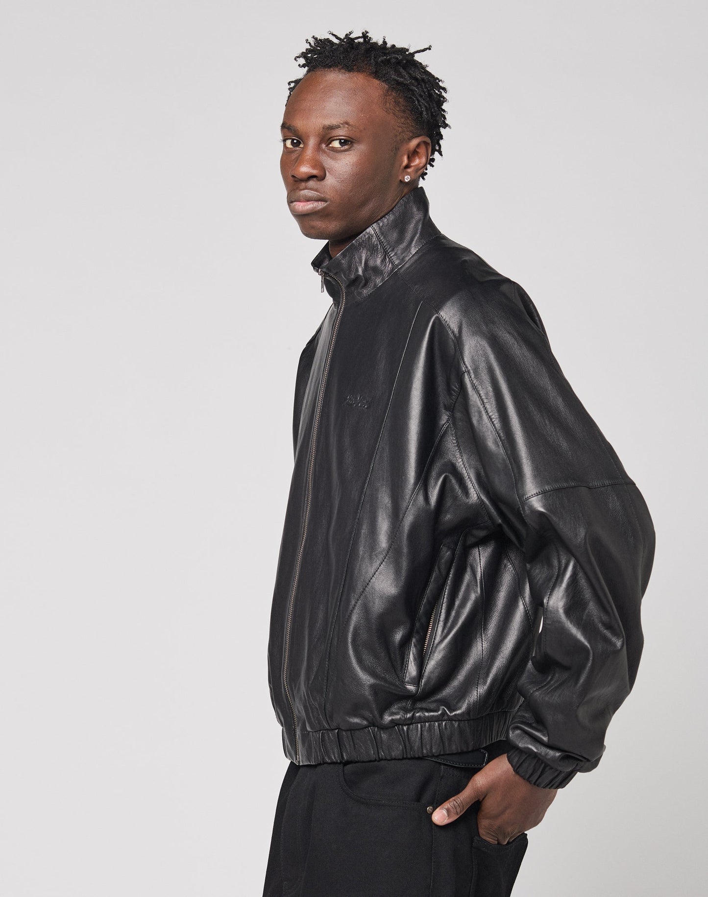 A man with short, textured hair wears the LIVE FAST DIE YOUNG Live Fast Leather Trackjacket with Stehkragen and dark pants, standing sideways and facing the camera against a plain light background.