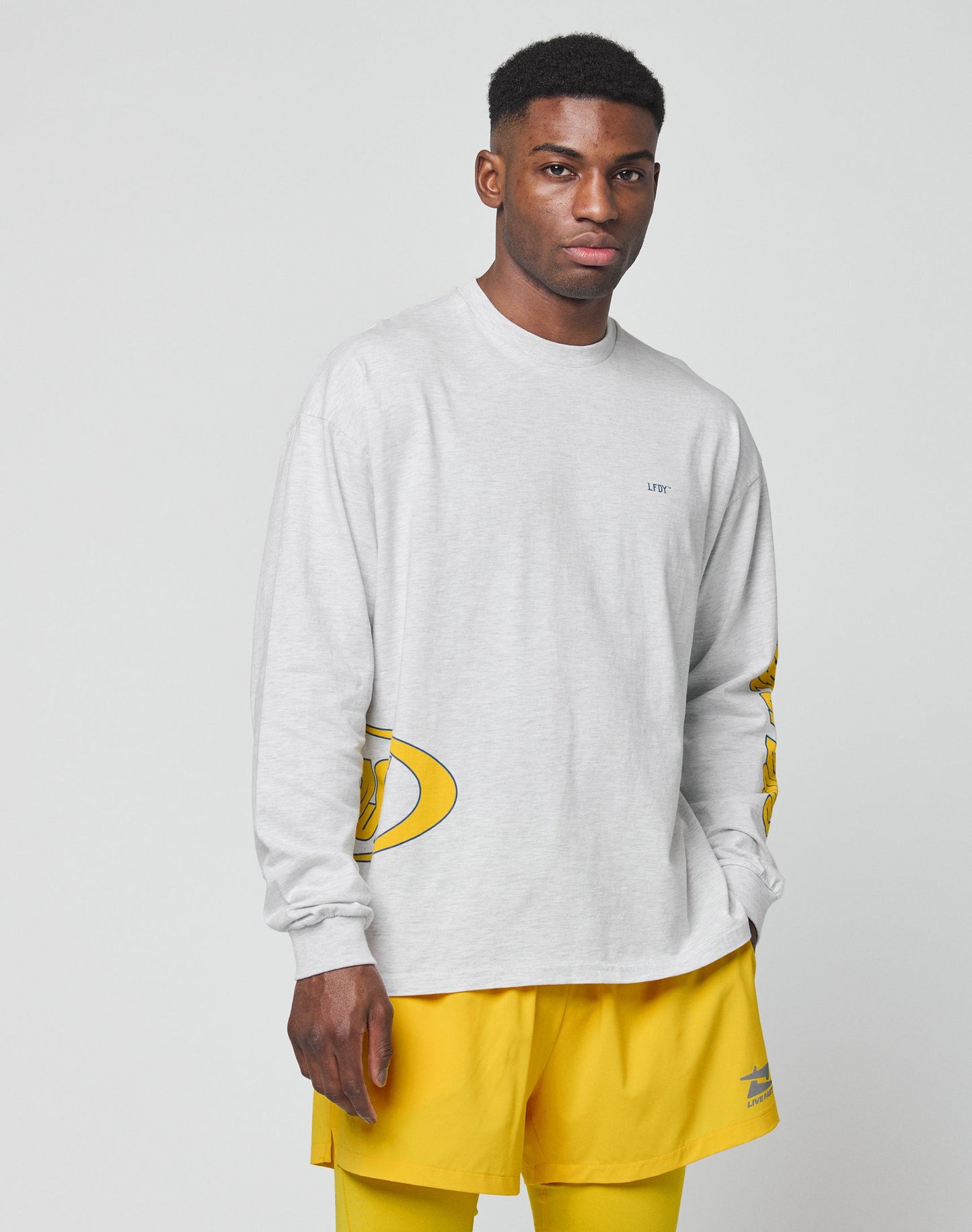 A man stands against a plain background wearing the LFDY Split Sphere Longsleeve by LIVE FAST DIE YOUNG, an oversize fit light gray shirt with small designs, paired with bright yellow shorts. He faces forward with a neutral expression, left hand by his side.