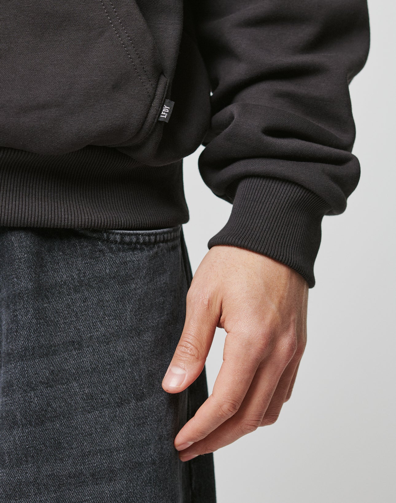 A close-up of a right hand resting by their side, wearing the LIVE FAST DIE YOUNG Live Fast Box Hooded sweatshirt and oversized dark denim jeans, set against a plain light background.