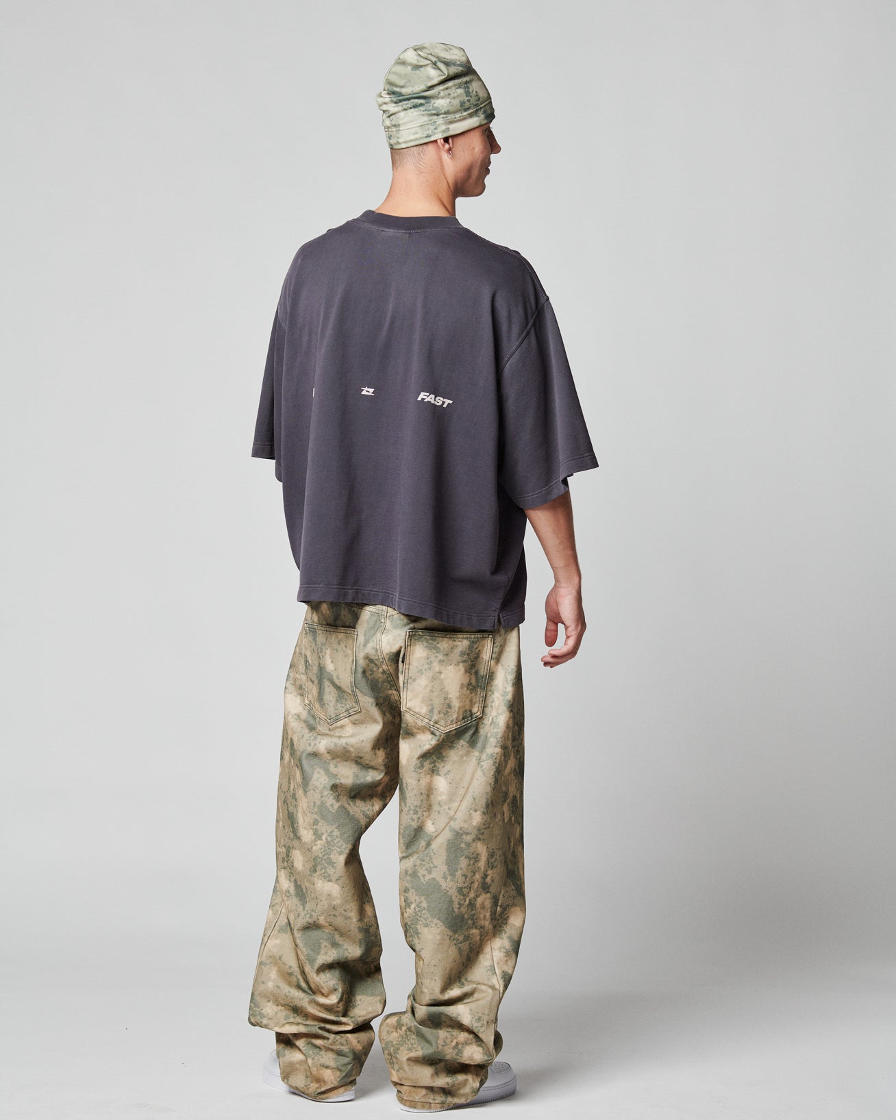 A person faces away, wearing the LIVE FAST DIE YOUNG Live Fast Cropped Oversized Tee with a bold backprint, paired with oversized camo pants, a matching camo beanie, and white shoes against a plain light gray background.