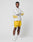 A man stands against a plain white background, confidently looking at the camera with arms crossed. He wears the LIVE FAST DIE YOUNG LFDY Split Sphere Longsleeve in light gray with yellow graphics, paired with yellow shorts, white socks, and sneakers.