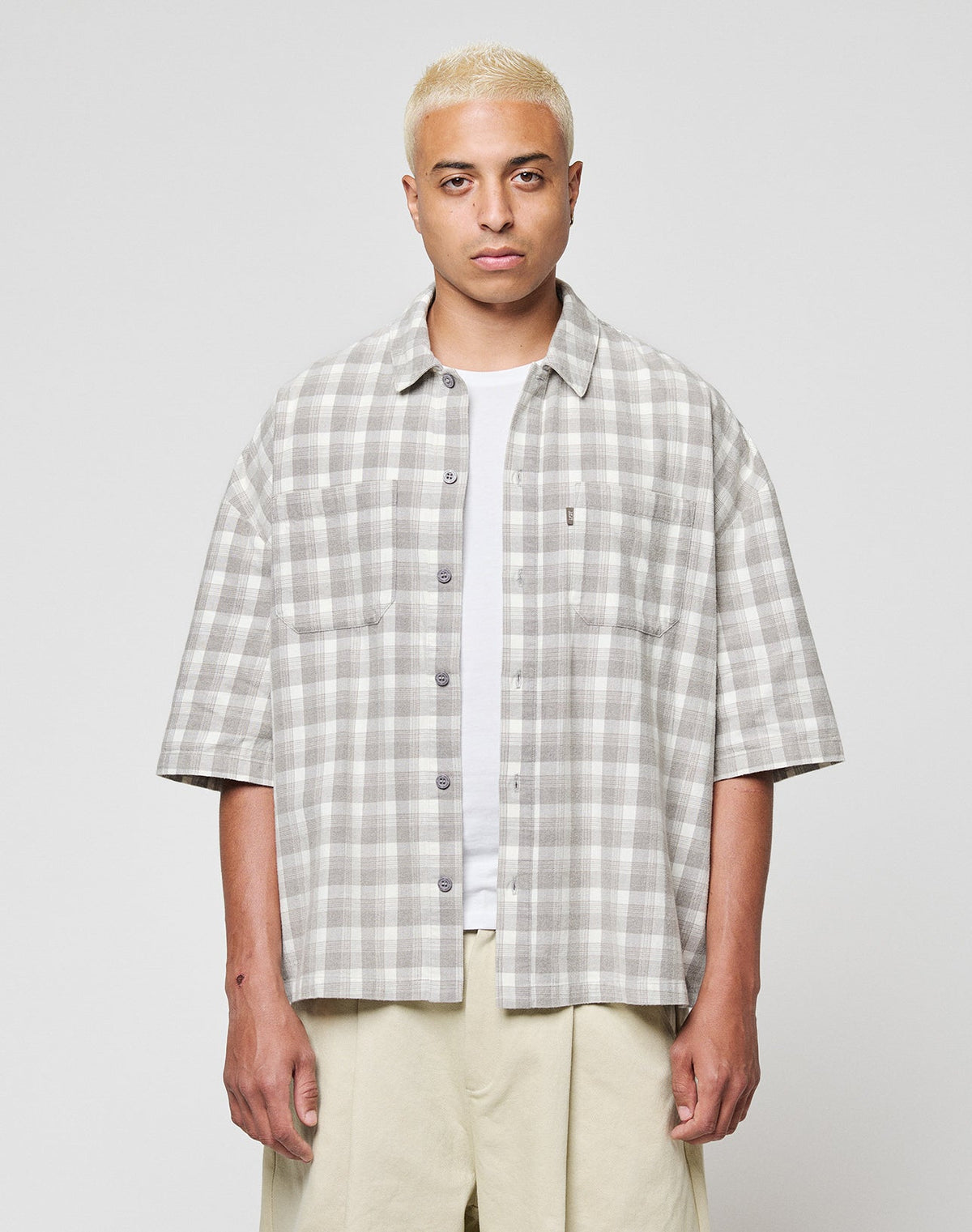 A person with short bleached blonde hair wears the LFDY Light Flannel by LIVE FAST DIE YOUNG in grey and white vintage wash over a white T-shirt and beige pants, standing against a plain light background. | model