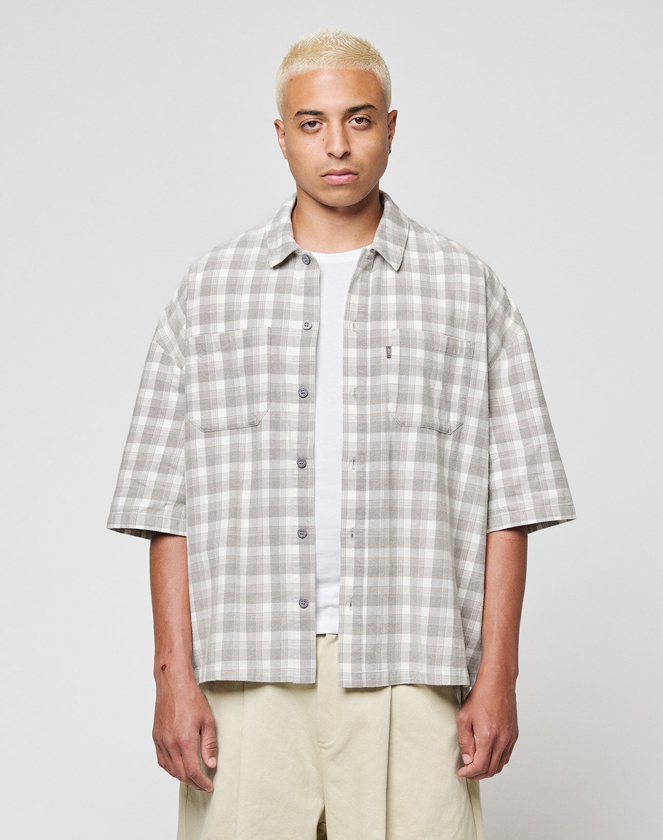 A person with short bleached blonde hair wears the LFDY Light Flannel by LIVE FAST DIE YOUNG in grey and white vintage wash over a white T-shirt and beige pants, standing against a plain light background.