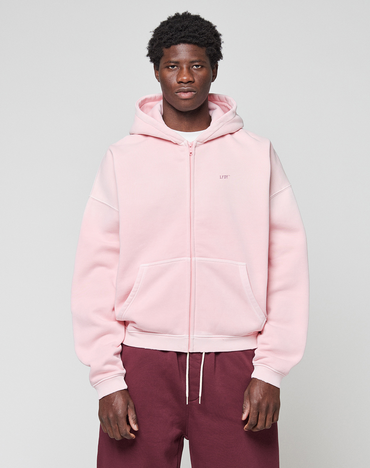 A person faces forward in a light pink LIVE FAST DIE YOUNG Basic Evidence Hooded Zip with an oversize fit and front pockets, paired with maroon pants, against a plain light gray background.