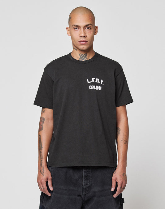 A person with a shaved head and visible tattoos wears the LFDY District Tee by LIVE FAST DIE YOUNG, featuring “L.F.D.Y. GMNH” front and back print, standing against a plain light gray background. | model