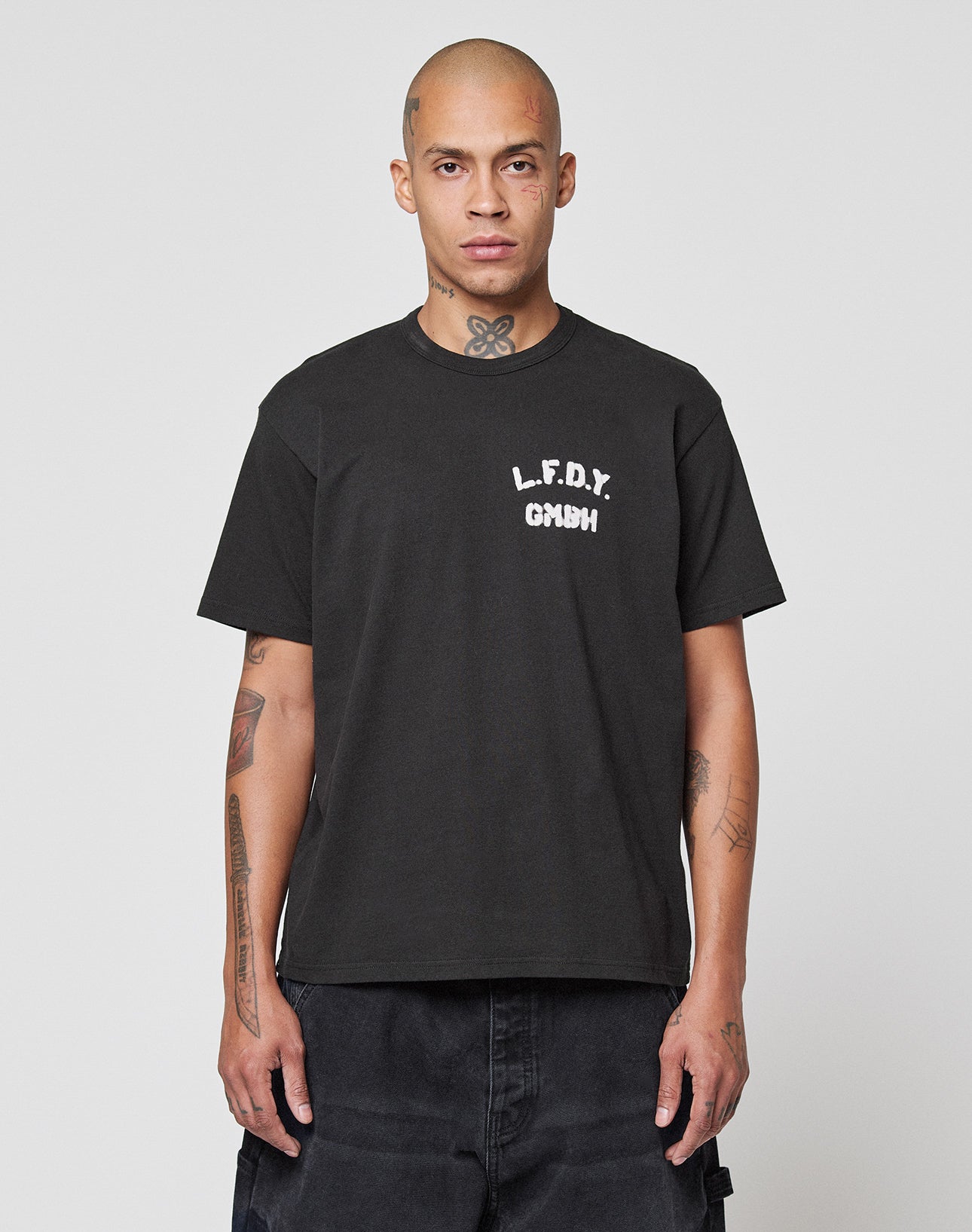 A person with a shaved head and visible tattoos wears the LFDY District Tee by LIVE FAST DIE YOUNG, featuring “L.F.D.Y. GMNH” front and back print, standing against a plain light gray background. | model