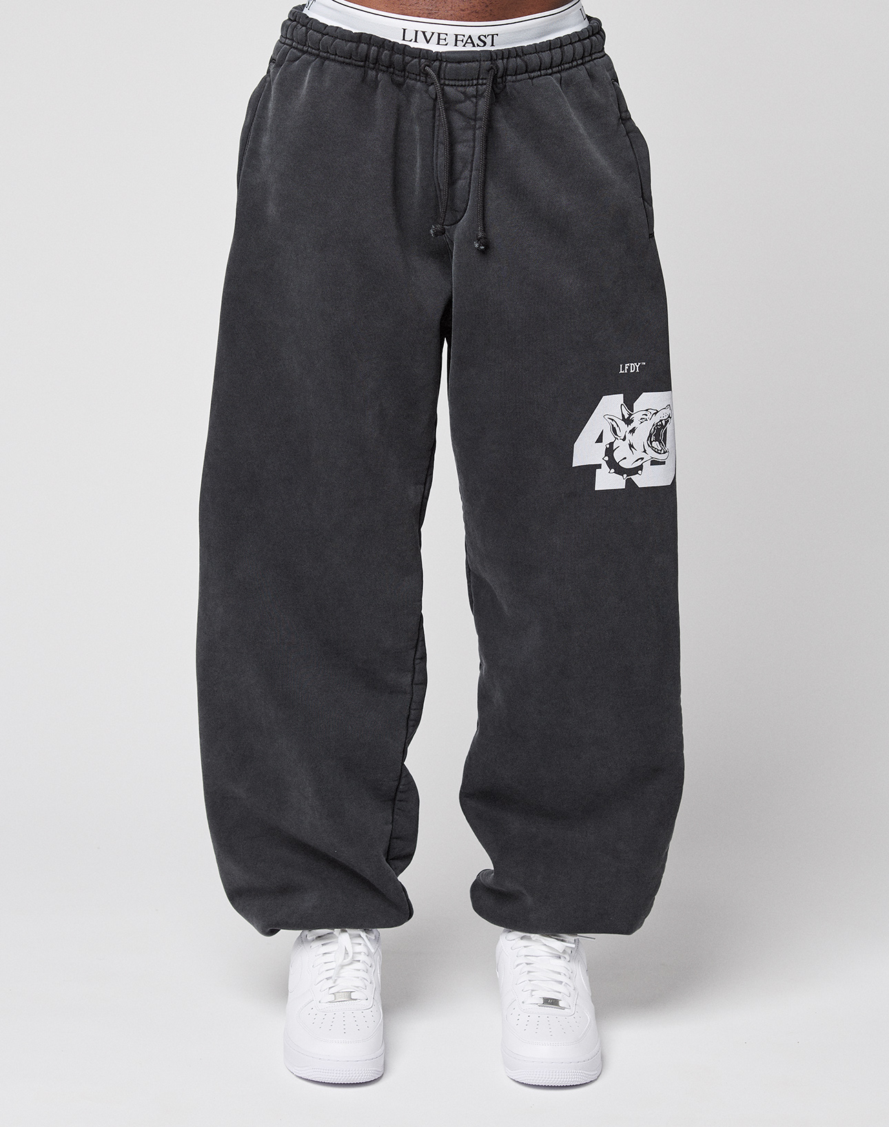 Someone wears the LFDY 40 Wide Cuff Jogger by LIVE FAST DIE YOUNG—oversized dark gray cotton sweatpants with a white tiger and "40" graphic on one leg, side pockets, paired with white sneakers and a visible LIVE FAST waistband.