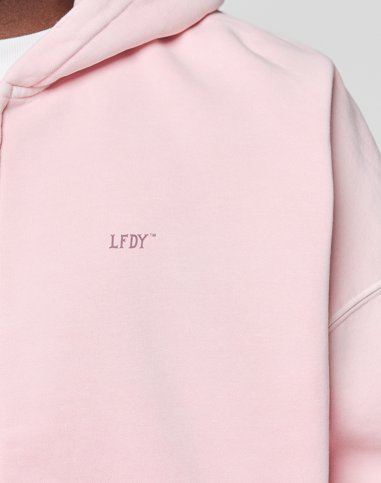 Close-up of a person wearing the light pink Basic Evidence Hooded Zip by LIVE FAST DIE YOUNG, featuring small purple LFDY-Branding embroidered on the chest. The hood and part of the shoulder are visible.