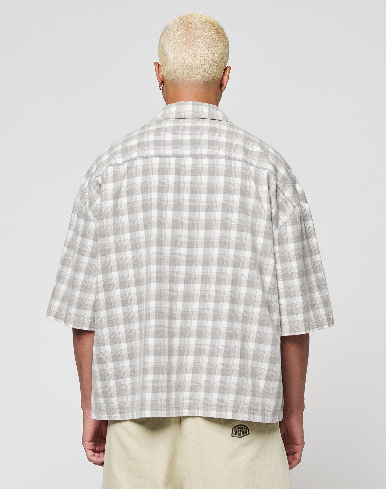 A person with short, bleached blonde hair is seen from the back wearing the LIVE FAST DIE YOUNG LFDY Light Flannel in grey and white plaid with a vintage wash and beige shorts, standing against a plain light background.