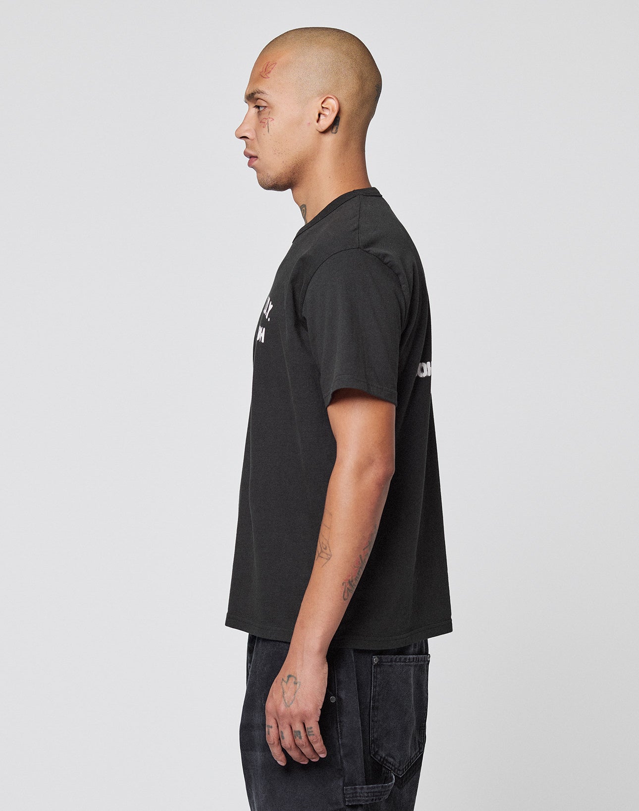 A person with a shaved head and visible tattoos stands in profile against a plain white background, wearing the black LIVE FAST DIE YOUNG LFDY District Tee and black pants.