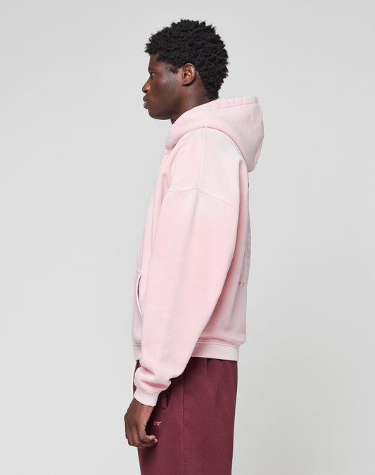 A person stands in profile against a plain light background, wearing the LIVE FAST DIE YOUNG Basic Evidence Hooded Zip in light pink with an oversize fit and maroon pants. Their hands are relaxed at their sides, hood down.