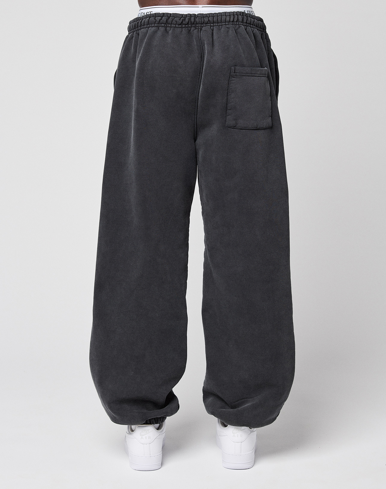 A person wearing LIVE FAST DIE YOUNG’s LFDY 40 Wide Cuff Jogger in dark gray with a white elastic waistband, side pockets, and a back pocket, paired with white sneakers, stands against a plain light background.