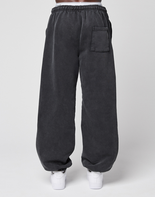 A person wearing LIVE FAST DIE YOUNG’s LFDY 40 Wide Cuff Jogger in dark gray with a white elastic waistband, side pockets, and a back pocket, paired with white sneakers, stands against a plain light background.
