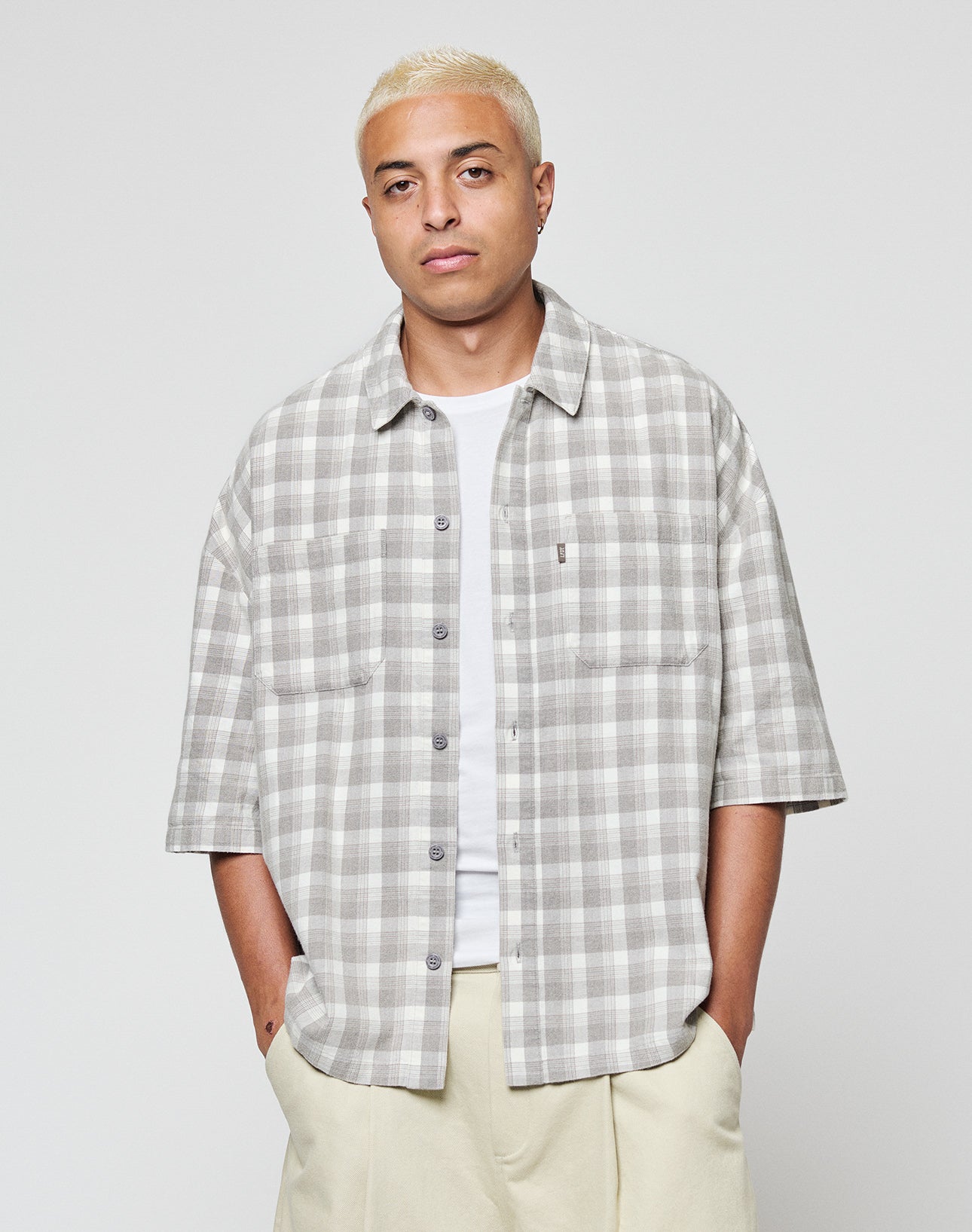 A person with short, bleached blond hair wears the LIVE FAST DIE YOUNG LFDY Light Flannel in gray and white plaid vintage wash over a white T-shirt and light-colored pants, standing against a plain light background.