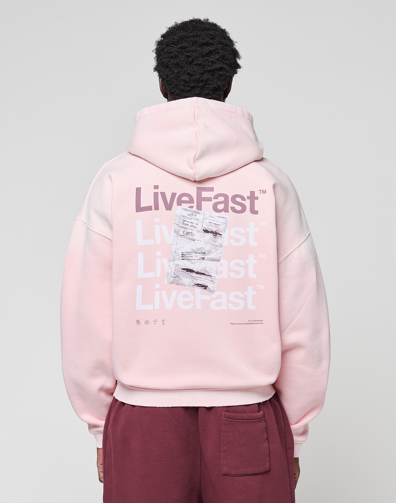 A person wears a LIVE FAST DIE YOUNG Basic Evidence Hooded Zip in oversize fit, light pink with white and purple "LiveFast" print and crumpled paper graphic on the back, paired with maroon pants. | model