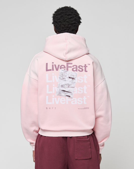 A person wears a LIVE FAST DIE YOUNG Basic Evidence Hooded Zip in oversize fit, light pink with white and purple "LiveFast" print and crumpled paper graphic on the back, paired with maroon pants. | model