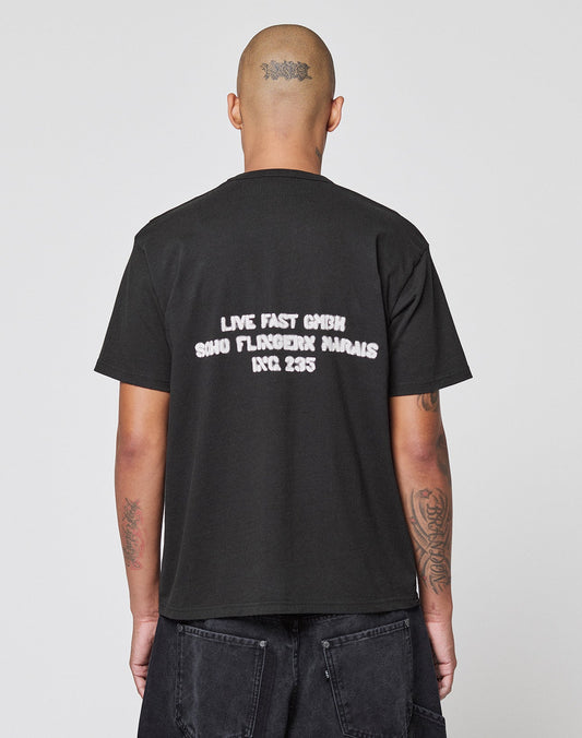 A tattooed, shaved-head individual wears the LFDY District Tee by LIVE FAST DIE YOUNG, a Regular Fit cotton shirt with front and back prints; the back reads: LIVE FAST GMBH SOHO FLAGSHIP MARAIS RCC 235. They stand before a plain background.