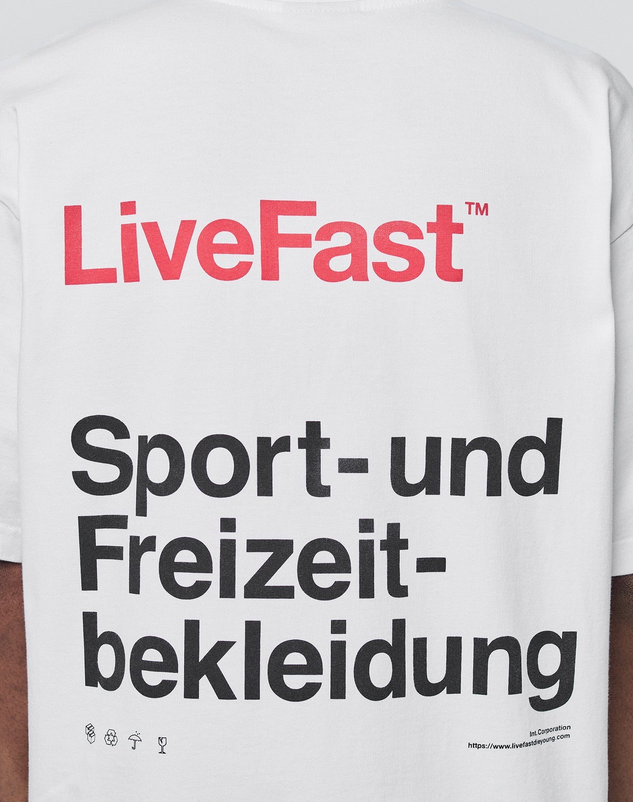 The Basic Trademark Tee by LIVE FAST DIE YOUNG is a white, 100% Baumwolle oversize shirt with bold red LiveFast™ text, a large black Sport- und Freizeitbekleidung backprint, small icons, and a website URL on the lower right.