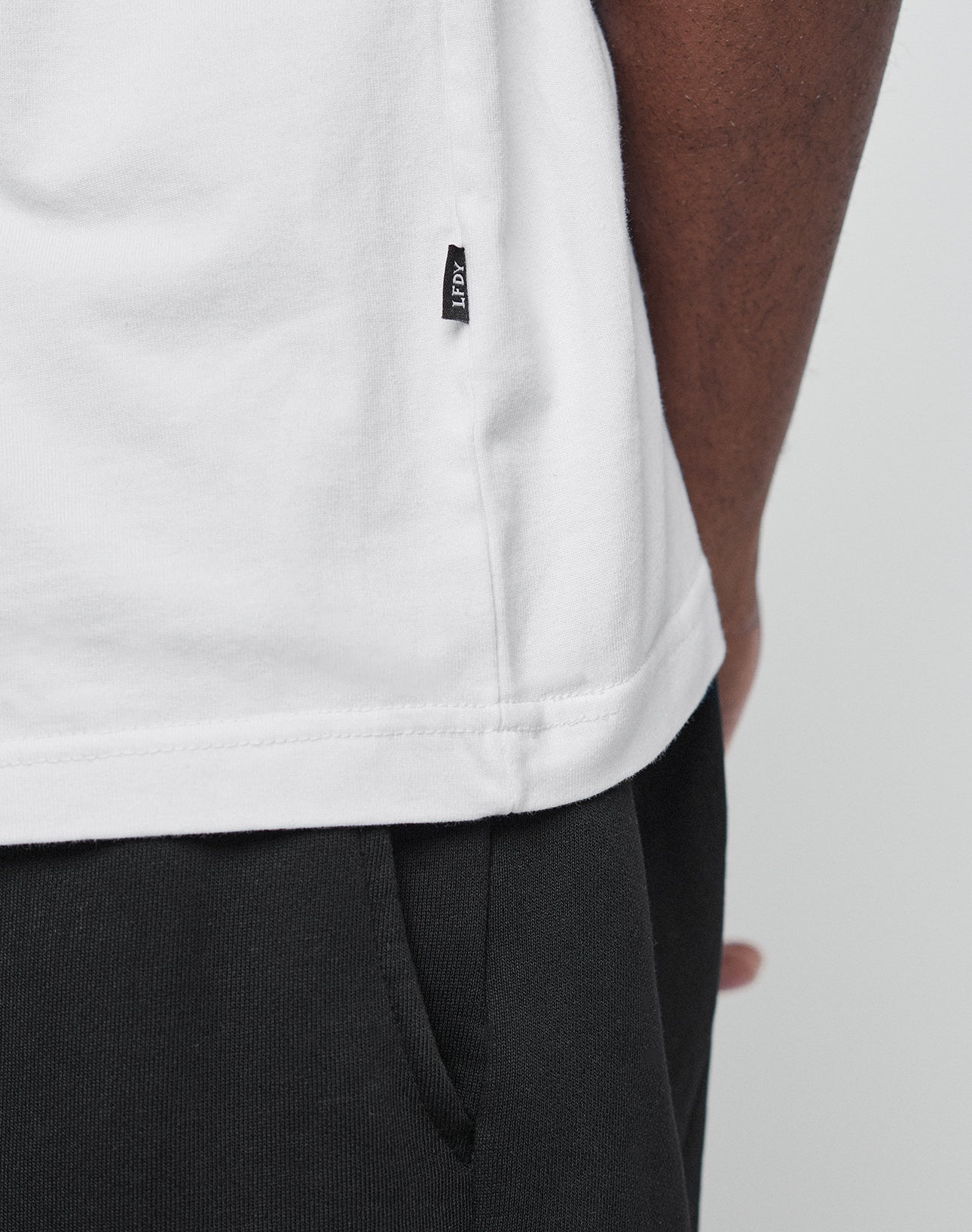Close-up of a person wearing the LIVE FAST DIE YOUNG Basic Trademark Tee—a white, 100% Baumwolle oversized t-shirt with a small black side tag. Paired with black pants and a hand near the pocket for a minimal look.