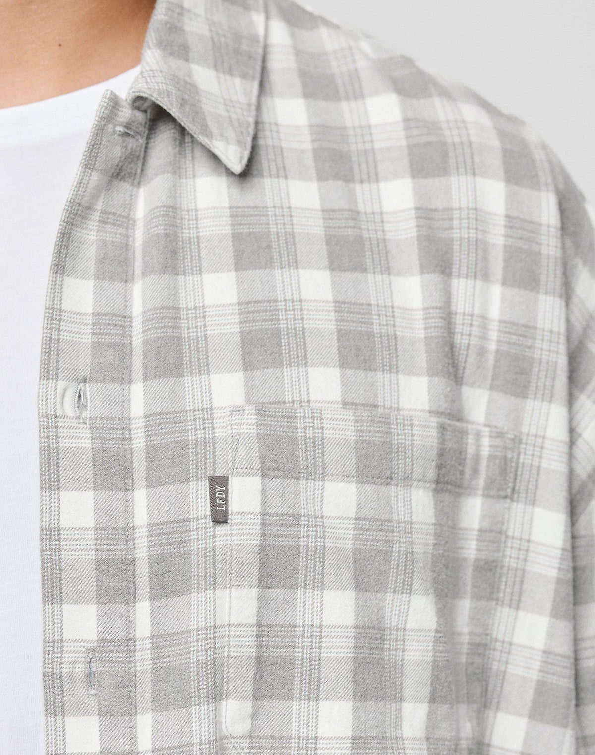 A close-up of a person in the LIVE FAST DIE YOUNG LFDY Light Flannel, a gray and white plaid oversize shirt with a chest pocket and small fabric tag, layered over a white tee; only their chest and neck are visible.