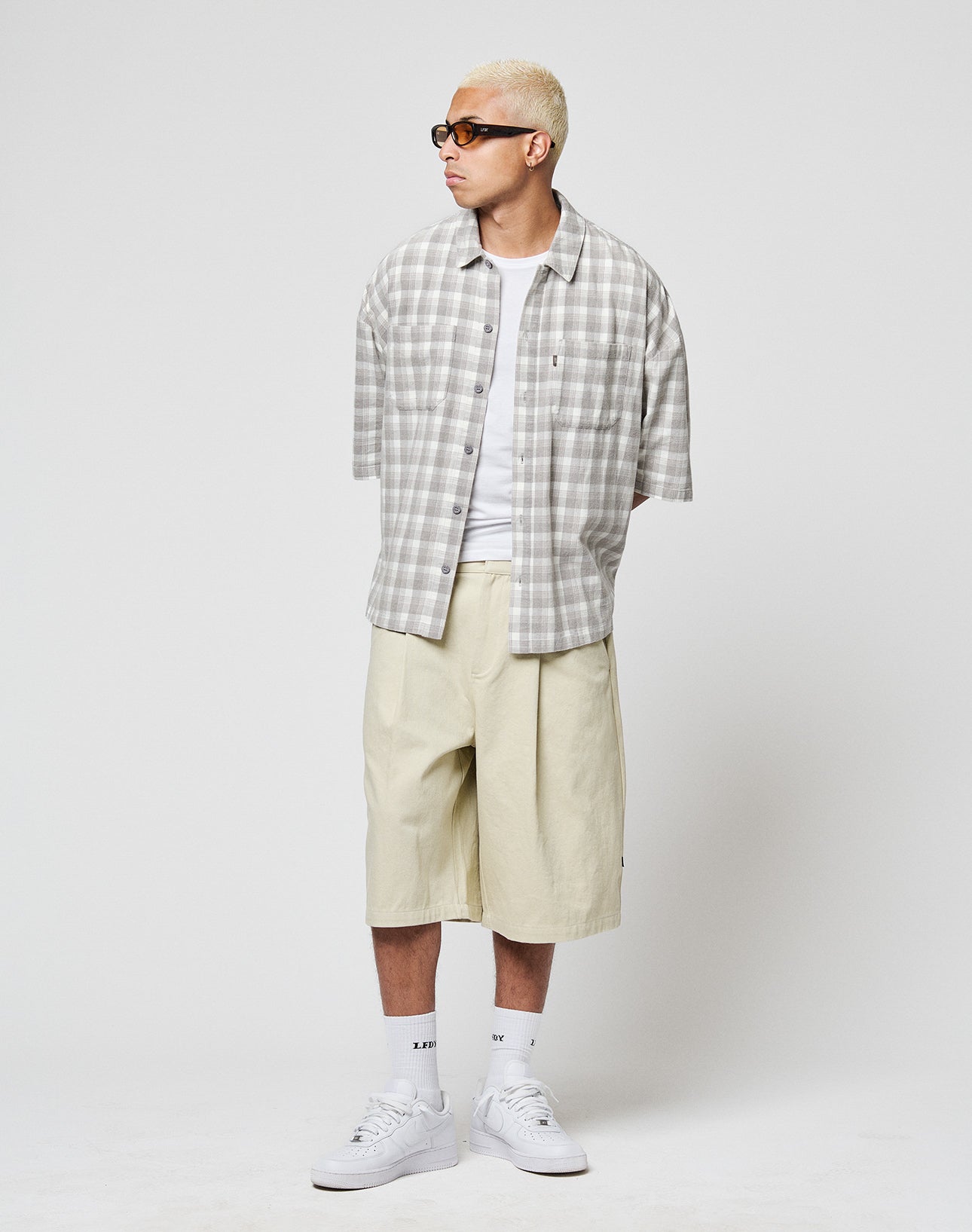 A man with short blond hair and sunglasses wears a LIVE FAST DIE YOUNG LFDY Light Flannel over a white tee, loose beige shorts, white socks, and sneakers while standing against a plain white background.