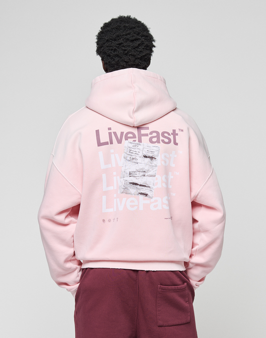 A person in an oversized light pink LIVE FAST DIE YOUNG Basic Evidence Hooded Zip, with repeated "LiveFast" text on the back, pairs it with maroon pants while standing facing away from the camera against a plain background.