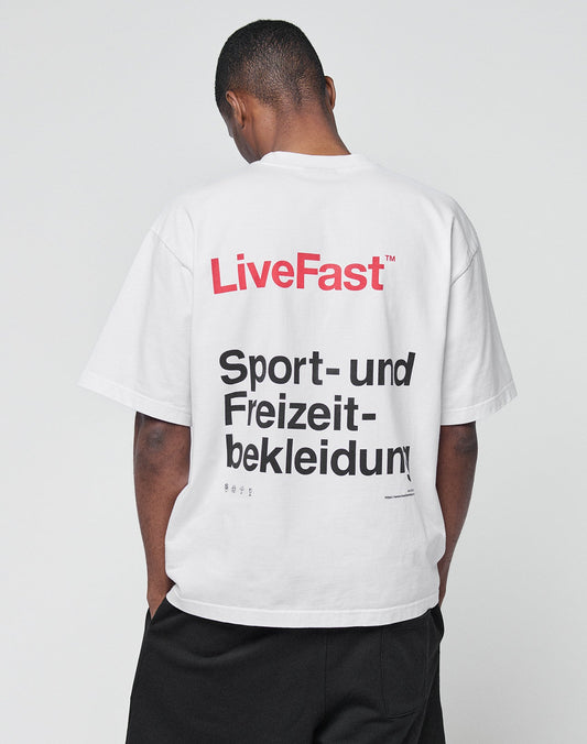 A person with short hair, back to camera, wears the LIVE FAST DIE YOUNG Basic Trademark Tee—an oversize white 100% Baumwolle T-shirt with bold LiveFast in red and Sport- und Freizeit-bekleidung in black on the back—paired with black pants.