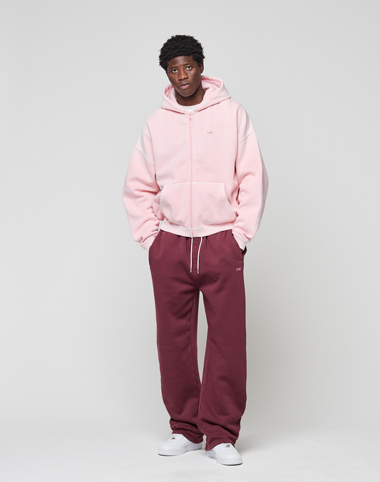 A person stands against a plain background wearing the LIVE FAST DIE YOUNG Basic Evidence Hooded Zip in light pink, paired with maroon sweatpants and white sneakers. Hands are in pockets, and they look relaxed.