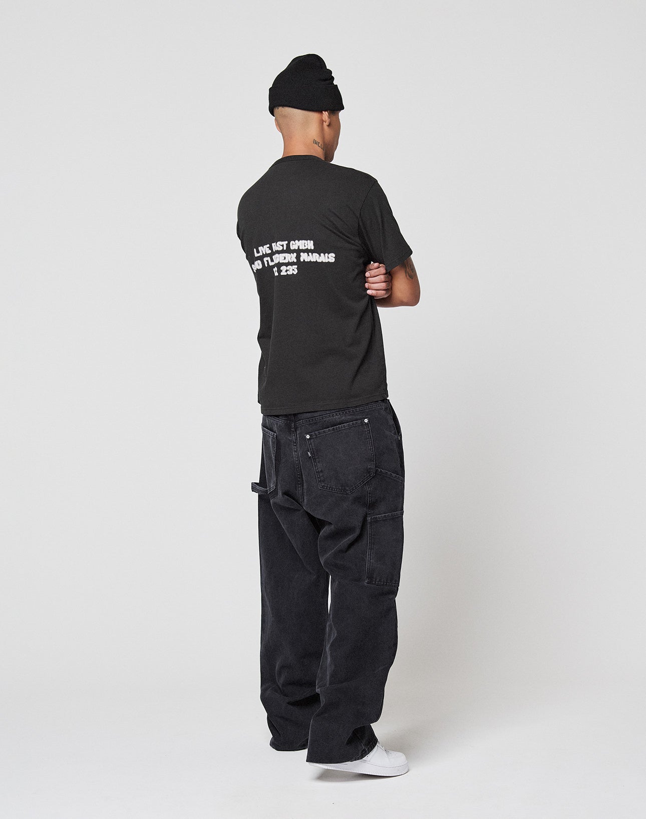 A person wears a LIVE FAST DIE YOUNG LFDY District Tee with front and back print, a black beanie, loose black jeans, and white sneakers, standing with arms crossed and facing away against a plain white background.