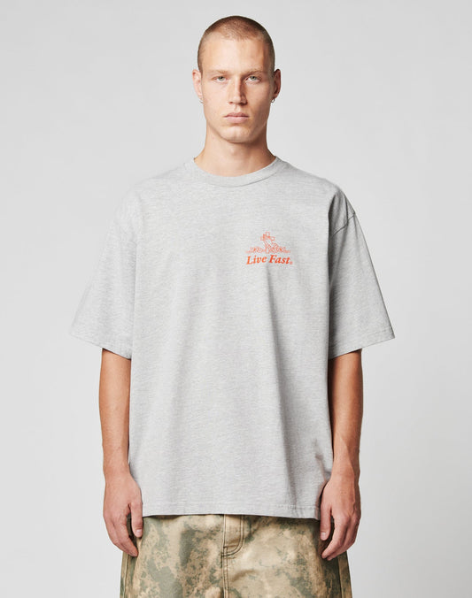 A person with short hair wears the LIVE FAST DIE YOUNG LFDY Praying Tee, an oversize light gray T-shirt with red Live Fast text, standing against a plain light gray background and wearing tan and green patterned pants.
