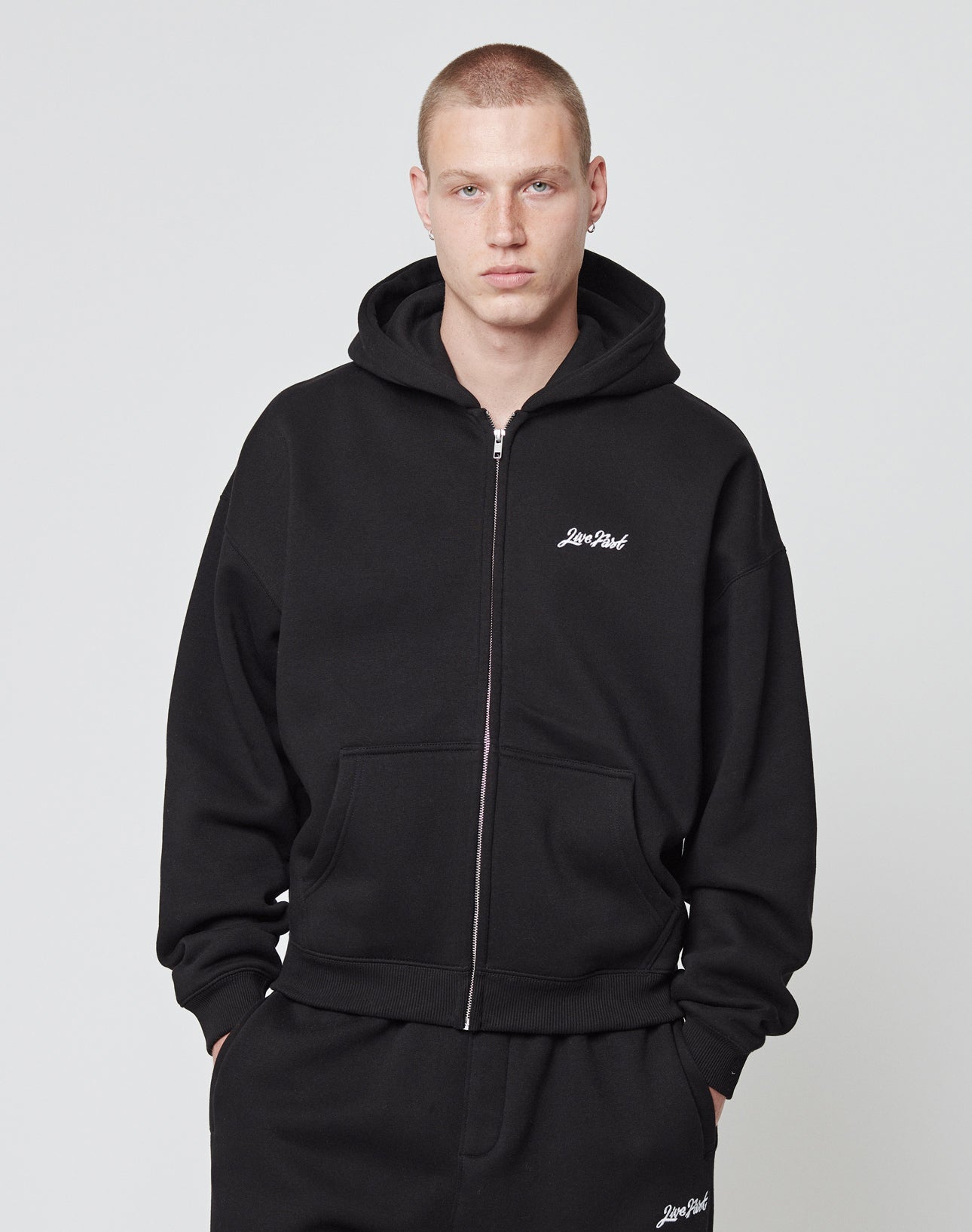 A person with short hair wears the LIVE FAST DIE YOUNG Basic Script Hooded Zip and matching black pants, both crafted from soft Baumwolle with subtle white logo-stitching on the left, standing against a plain light background with hands in pockets.