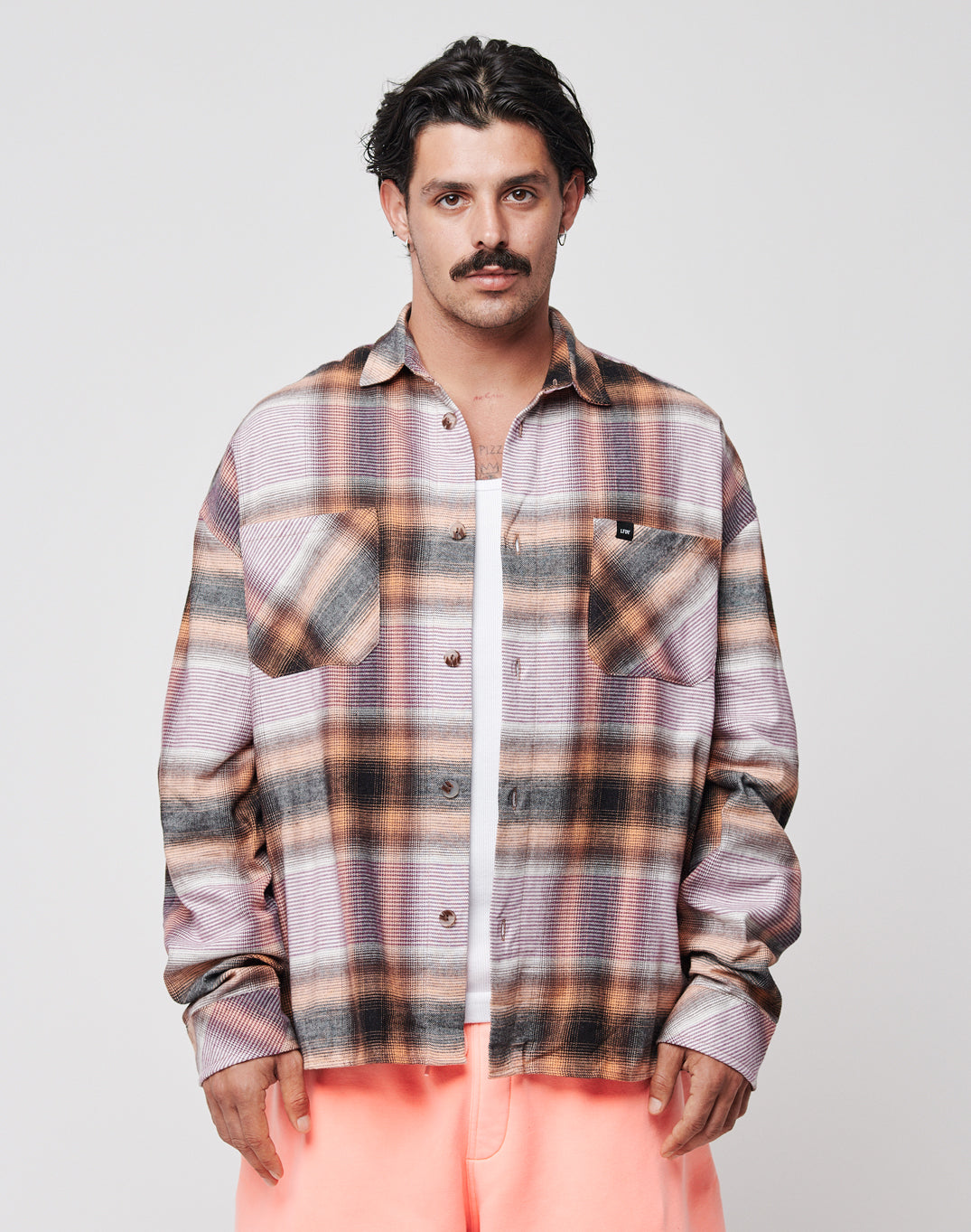 A dark-haired man with a mustache wears the LIVE FAST DIE YOUNG Loose Summer Flannel—an oversize pink, brown, and gray plaid shirt—over a white tee and light pink pants, standing against a plain light background.