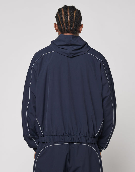 A person with braided hair, seen from the back, wears the LIVE FAST DIE YOUNG Live Fast Trackjacket with white piping, elastic cuffs, zip pockets, and matching pants against a simple light background.