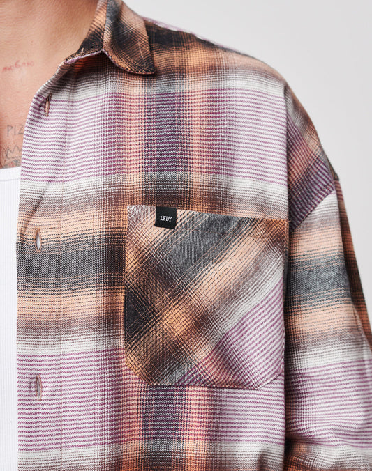 A close-up of a person wearing LIVE FAST DIE YOUNG’s Loose Summer Flannel—a 100% cotton plaid shirt in pink, orange, and black with a chest pocket LFDTY label—layered over a white undershirt.