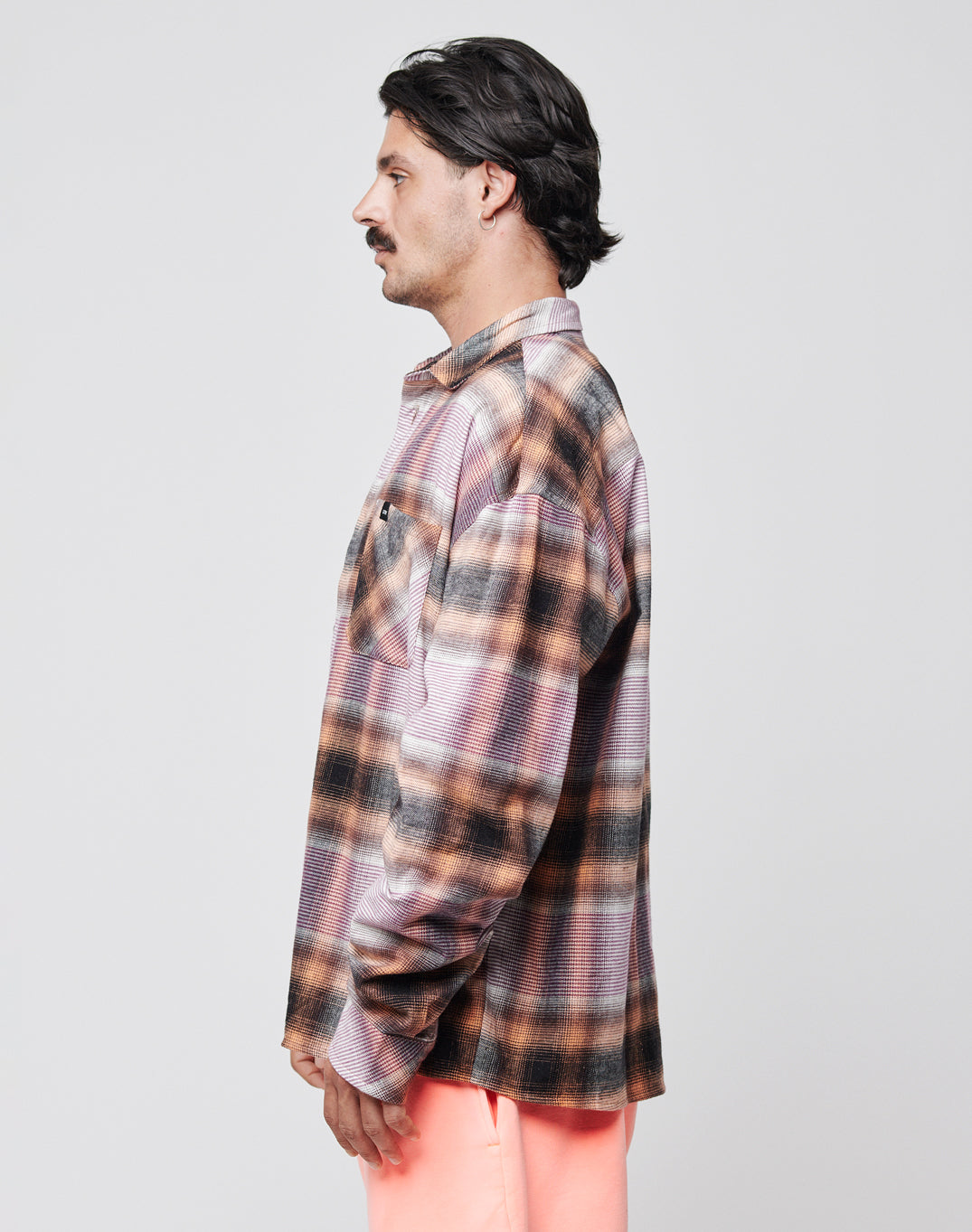 A man with dark hair and a mustache stands in profile, wearing the LIVE FAST DIE YOUNG Loose Summer Flannel in plaid brown, orange, and black tones with light pink pants against a plain light gray background.