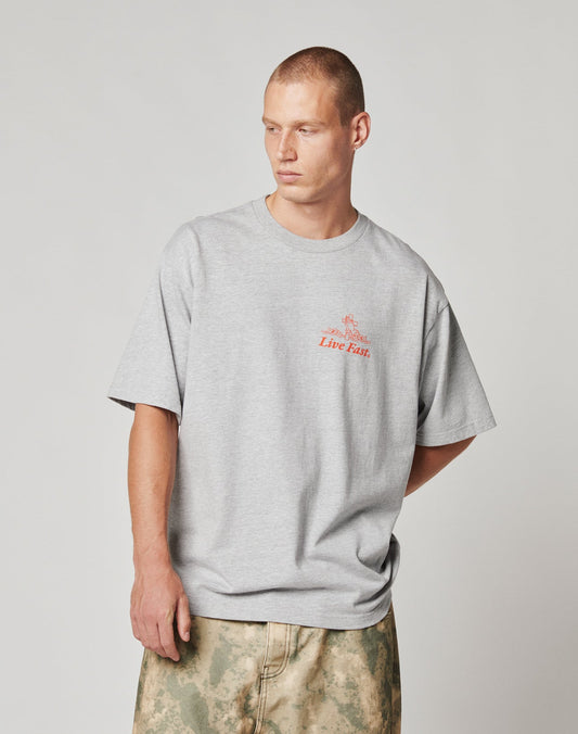 A man with short hair wears the LIVE FAST DIE YOUNG LFDY Praying Tee, an oversize fit gray T-shirt with red chest text, paired with light green camo pants, standing against a plain light background.