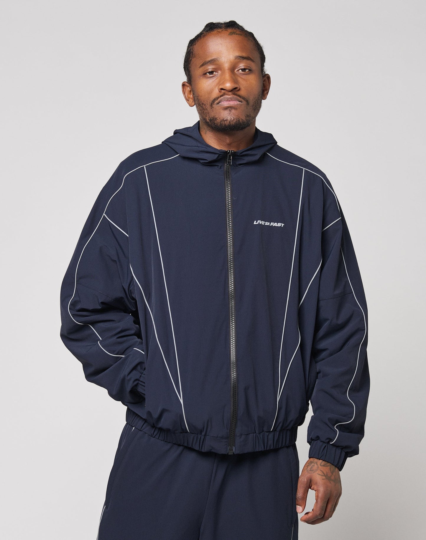 A man wears the LIVE FAST DIE YOUNG Live Fast Trackjacket in navy blue with reflektierender Druck and elastische Bündchen, standing against a plain light background, looking at the camera with a neutral expression and one hand in his pocket.