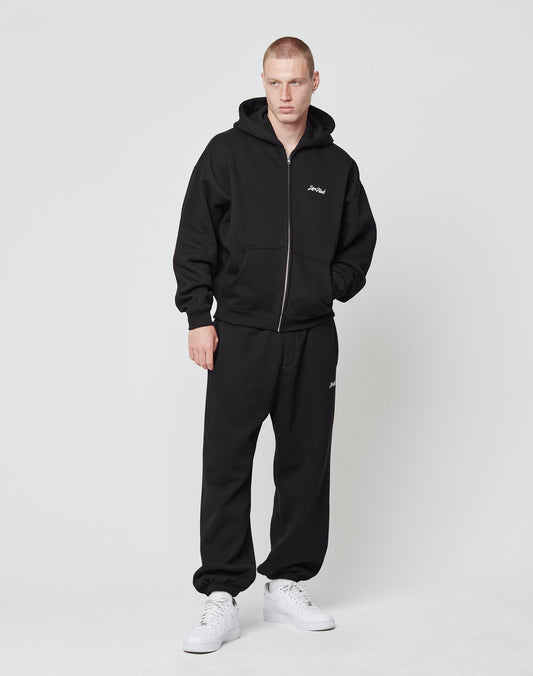 A person stands against a plain background wearing LIVE FAST DIE YOUNG’s Loose Jogger—a loose fit black hoodie in weicher Baumwolle with a small white chest logo, paired with matching black sweatpants and white sneakers.