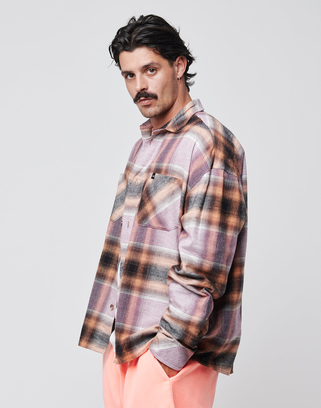 A man with dark hair and a mustache wears the LIVE FAST DIE YOUNG Loose Summer Flannel—a vintage wash, oversize plaid shirt in purple, orange, and black—paired with light pink pants against a plain white background.