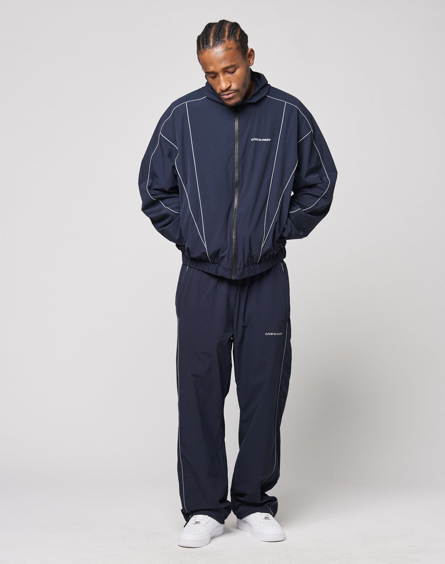 A man stands against a plain light background, wearing LIVE FAST DIE YOUNGs Live Fast Trackpants in navy with elastic waistband, white piping, and reflective logo print. He pairs them with white sneakers, hands in pockets, looking down.