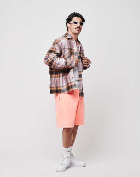 A man wearing white sunglasses, a LIVE FAST DIE YOUNG Loose Summer Flannel in vintage wash over a white tee, peach shorts, white socks, and sneakers poses against a plain white background. The shirt is made from 100% Baumwolle.