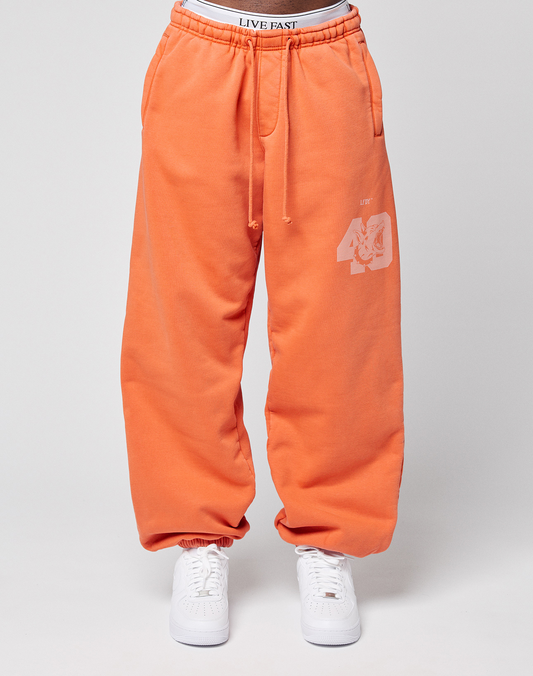 A person wears LFDY 40 Wide Cuff Jogger by LIVE FAST DIE YOUNG—bright orange joggers with a white “40” and tiger face graphic on the left thigh, oversize fit, elastic waistband with “LIVE FAST” text—paired with white sneakers against a light gray background.