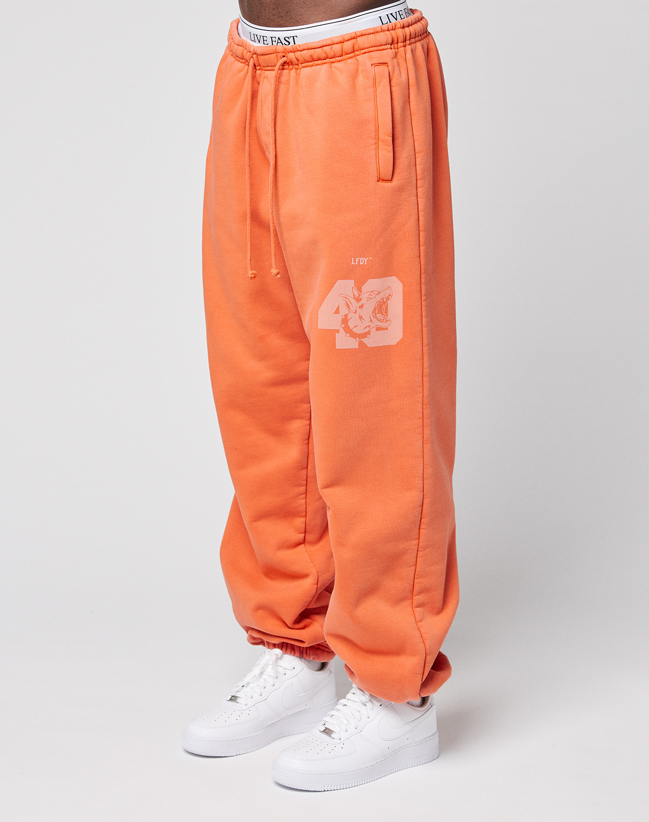 A person wears the LFDY 40 Wide Cuff Jogger by LIVE FAST DIE YOUNG: loose-fitting orange sweatpants with Oversize Fit, white "40" print on the leg, side pockets, an elastic waistband with "LIVE FAST" text, paired with white sneakers. Plain light background.