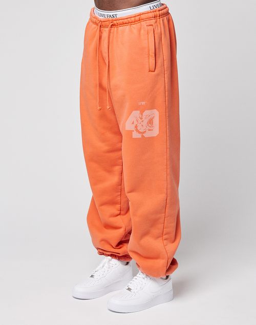 A person wears the LFDY 40 Wide Cuff Jogger by LIVE FAST DIE YOUNG: loose-fitting orange sweatpants with Oversize Fit, white "40" print on the leg, side pockets, an elastic waistband with "LIVE FAST" text, paired with white sneakers. Plain light background.