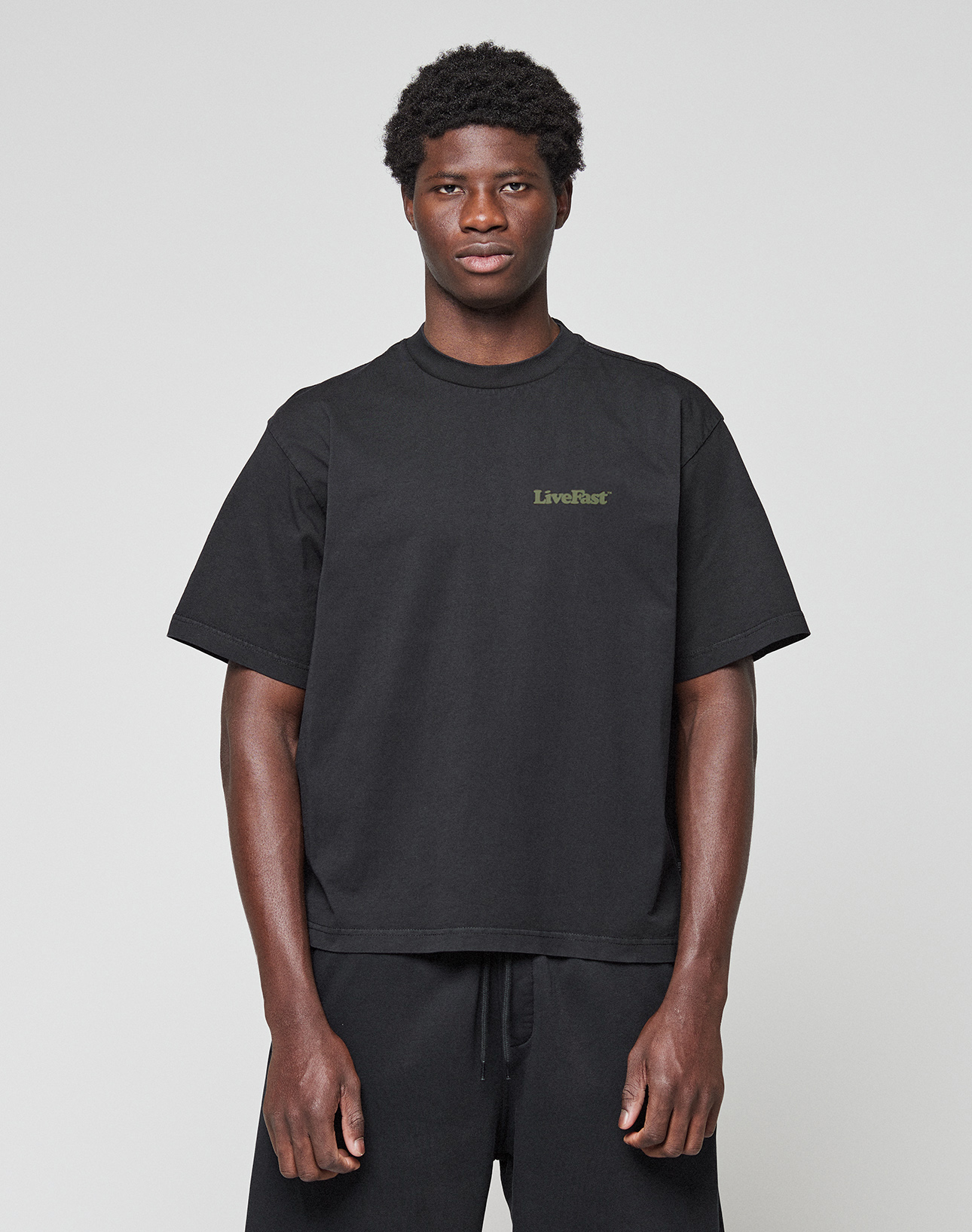 A person faces forward, wearing the LIVE FAST DIE YOUNG Basic 210 Tee—an oversized black short-sleeve shirt made from 100% cotton with "LiveFast" in small green letters on the left chest—and black pants, against a plain light gray background.
