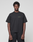 A person faces forward, wearing the LIVE FAST DIE YOUNG Basic 210 Tee—an oversized black short-sleeve shirt made from 100% cotton with "LiveFast" in small green letters on the left chest—and black pants, against a plain light gray background.