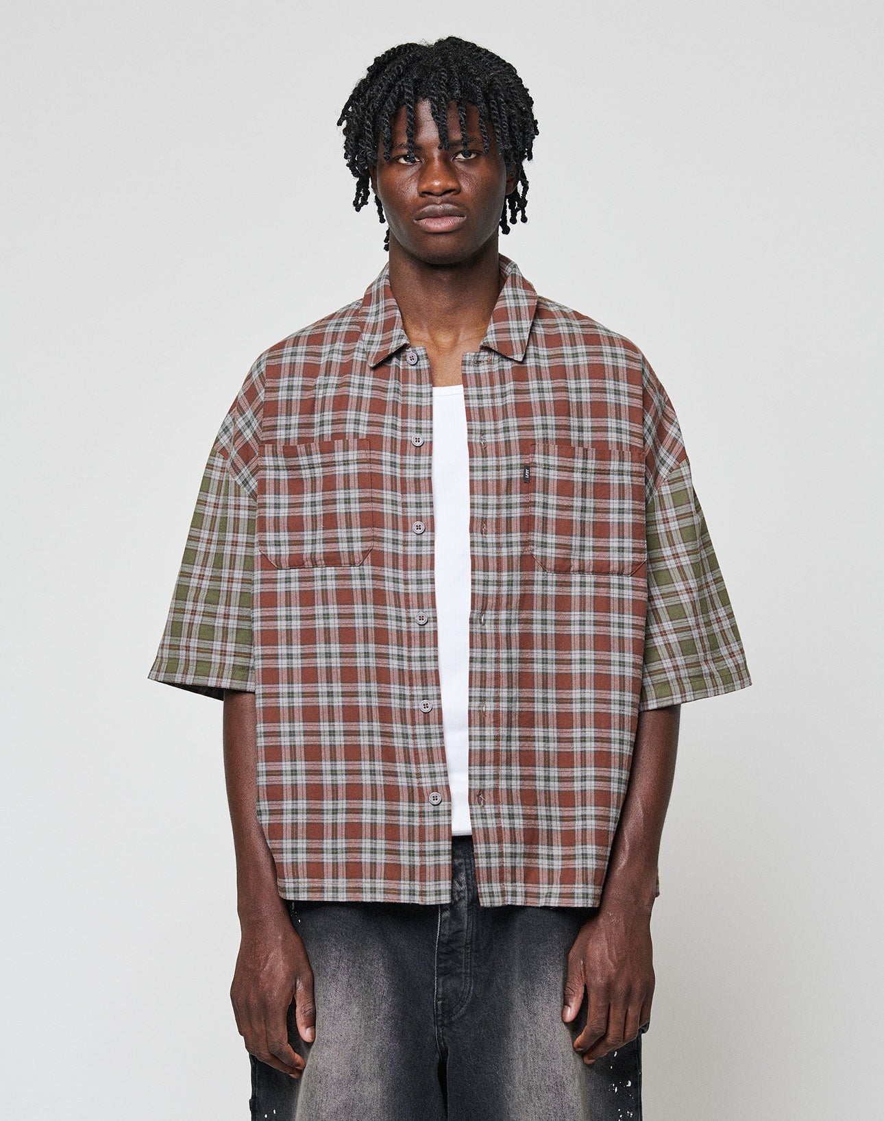 A young man with short dreadlocks wears the LIVE FAST DIE YOUNG LFDY Light Flannel—an oversize fit, short-sleeve, plaid button-up in brown, green, and grey—over a white tee and dark jeans against a plain light background. | model